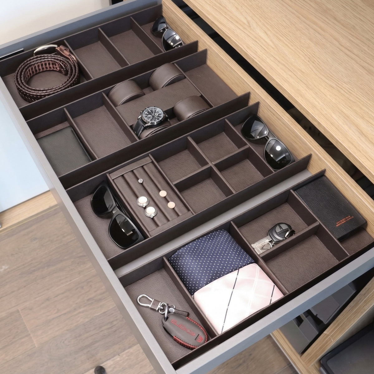 An overhead view of an open wardrobe drawer fitted with multiple brown modular organizer trays. The organized compartments contain various accessories including a coiled belt, sunglasses, a wristwatch on a cushion, cufflinks on a padded roll, folded ties, a wallet, and keys.