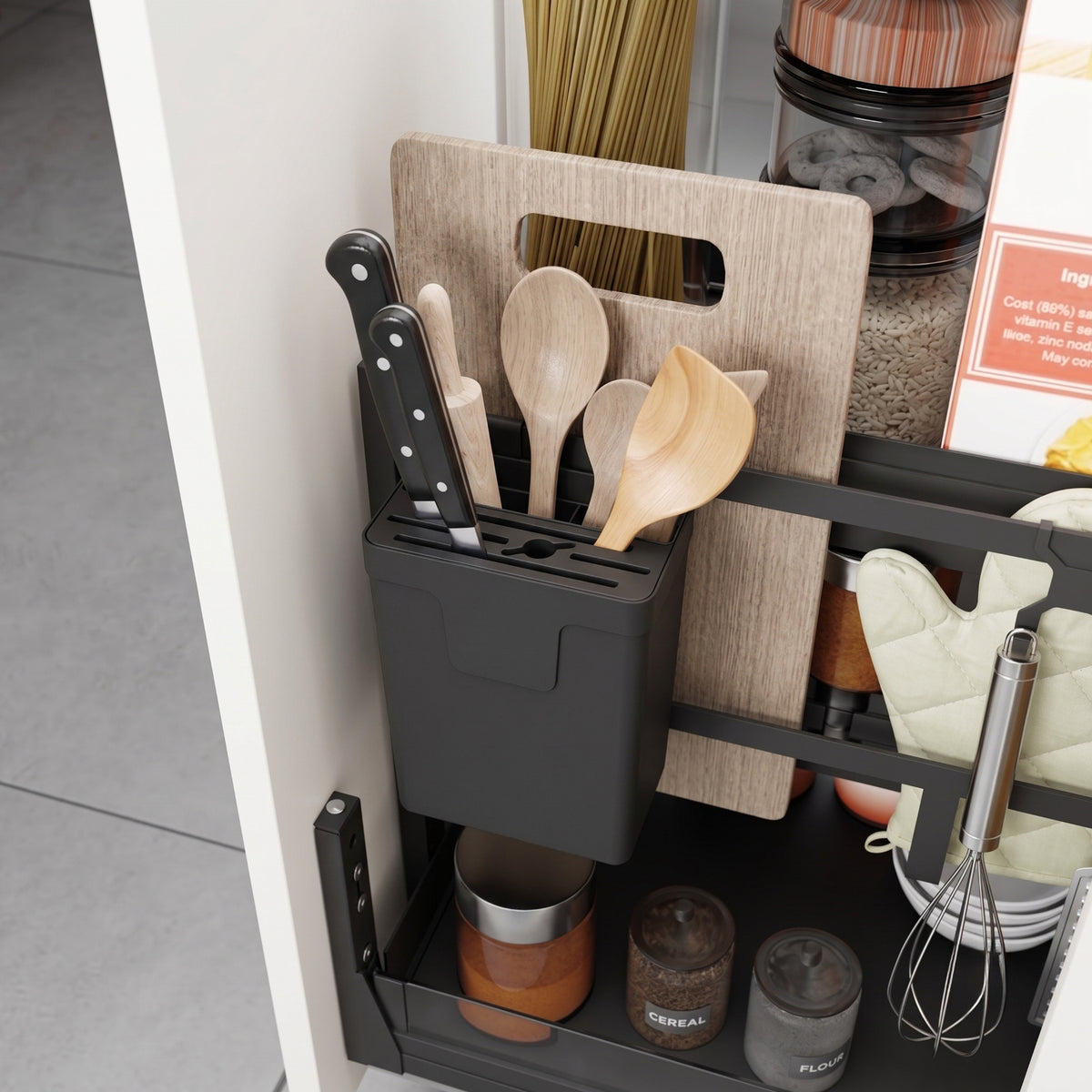 A close-up photograph of a modern white kitchen cabinet featuring an extended black pull-out storage system. The upper level contains a black utensil organizer with knives, wooden spoons, and wooden spatulas. A light-wood cutting board is stored behind the organizer. Various items are visible on the pull-out shelves, including bundles of pasta, jars of cereal and granola, labeled spice jars for 'CEREAL' and 'FLOUR', a white oven mitt, a wire whisk, and white dishes. The background shows grey tile flooring.