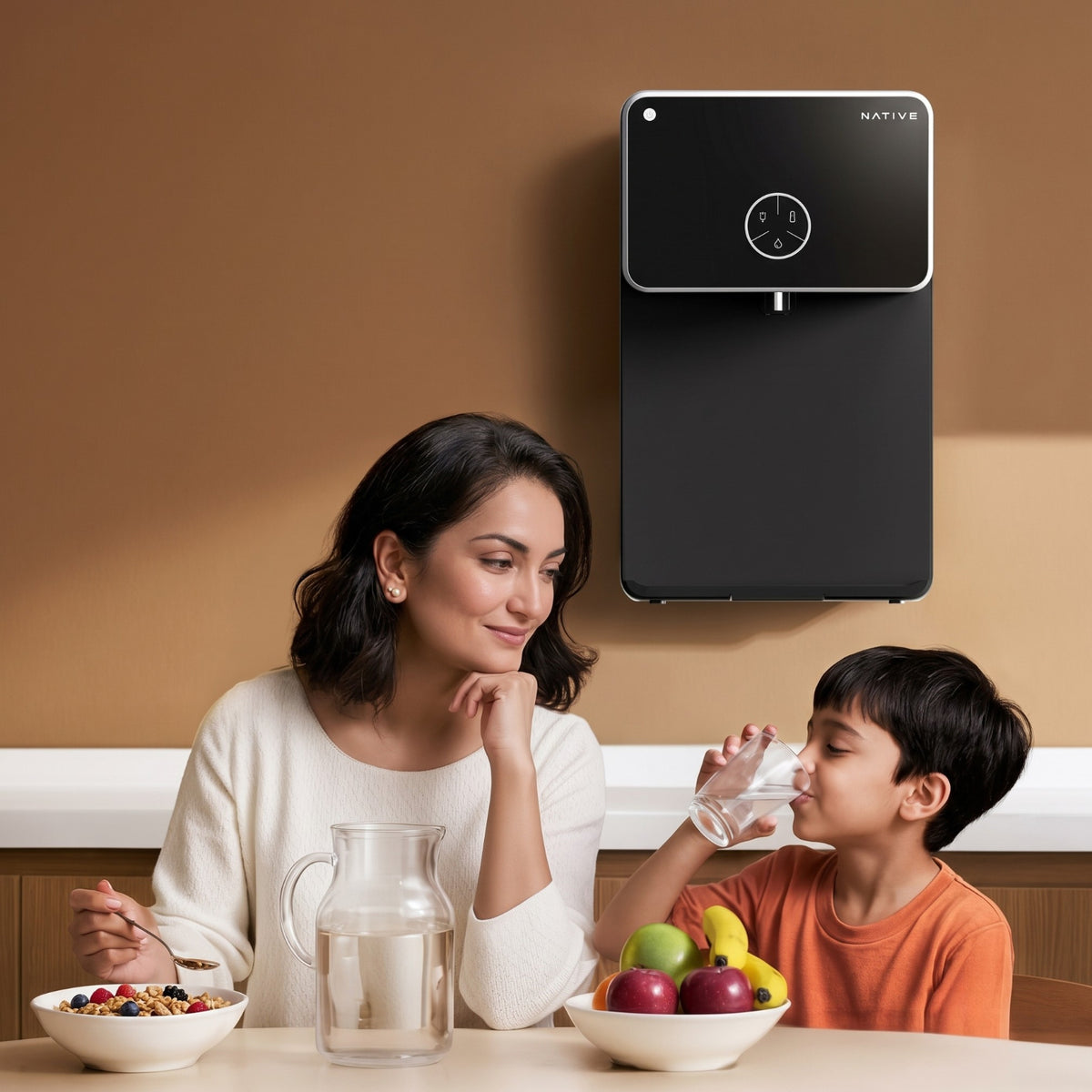 A mother and child sitting at a kitchen table drinking water and eating fruit, with the Native water purifier mounted on the wall behind them