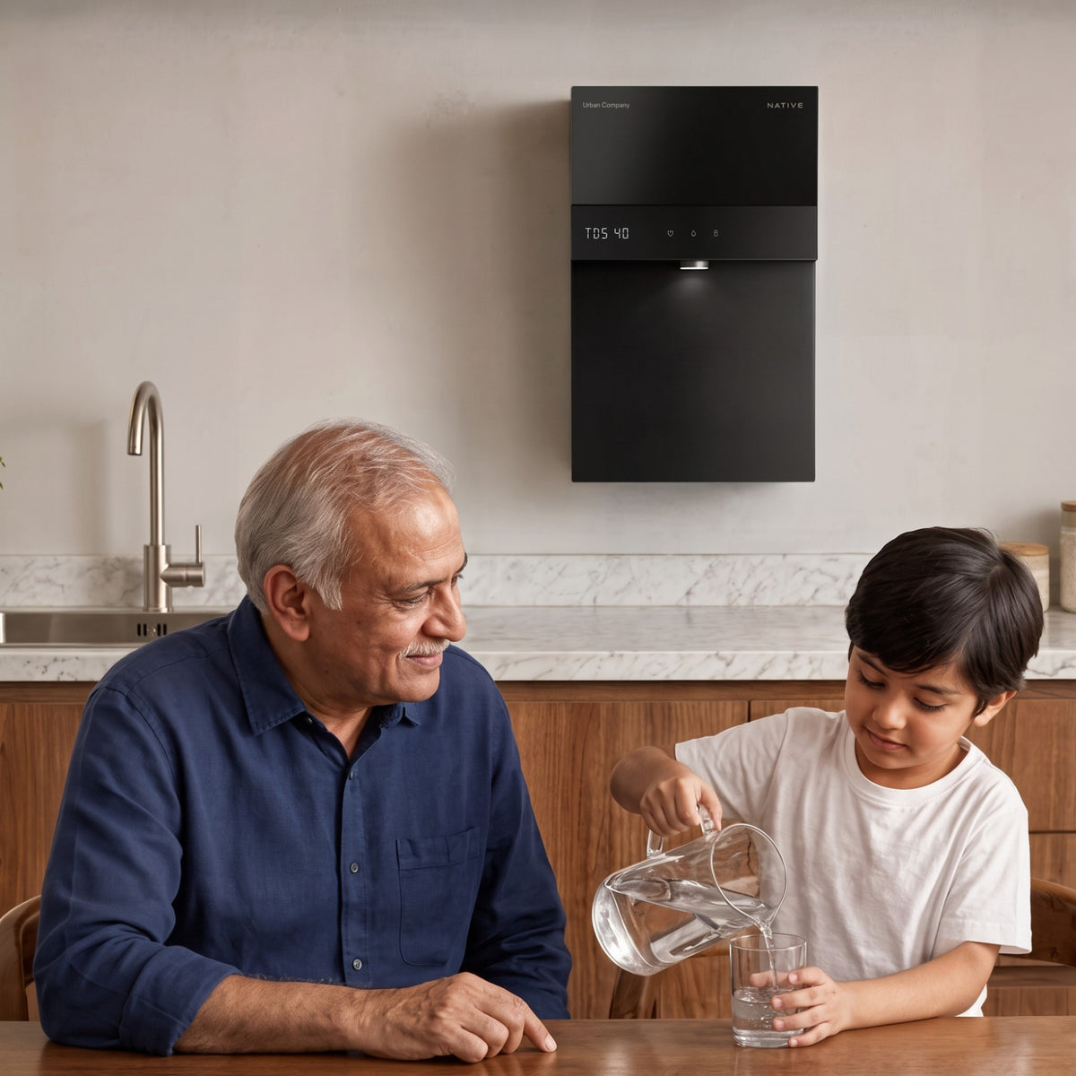 A young boy pouring water from a glass pitcher into a cup while sitting at a kitchen table with an older man, with the black Urban Company Native water purifier mounted on the wall behind them