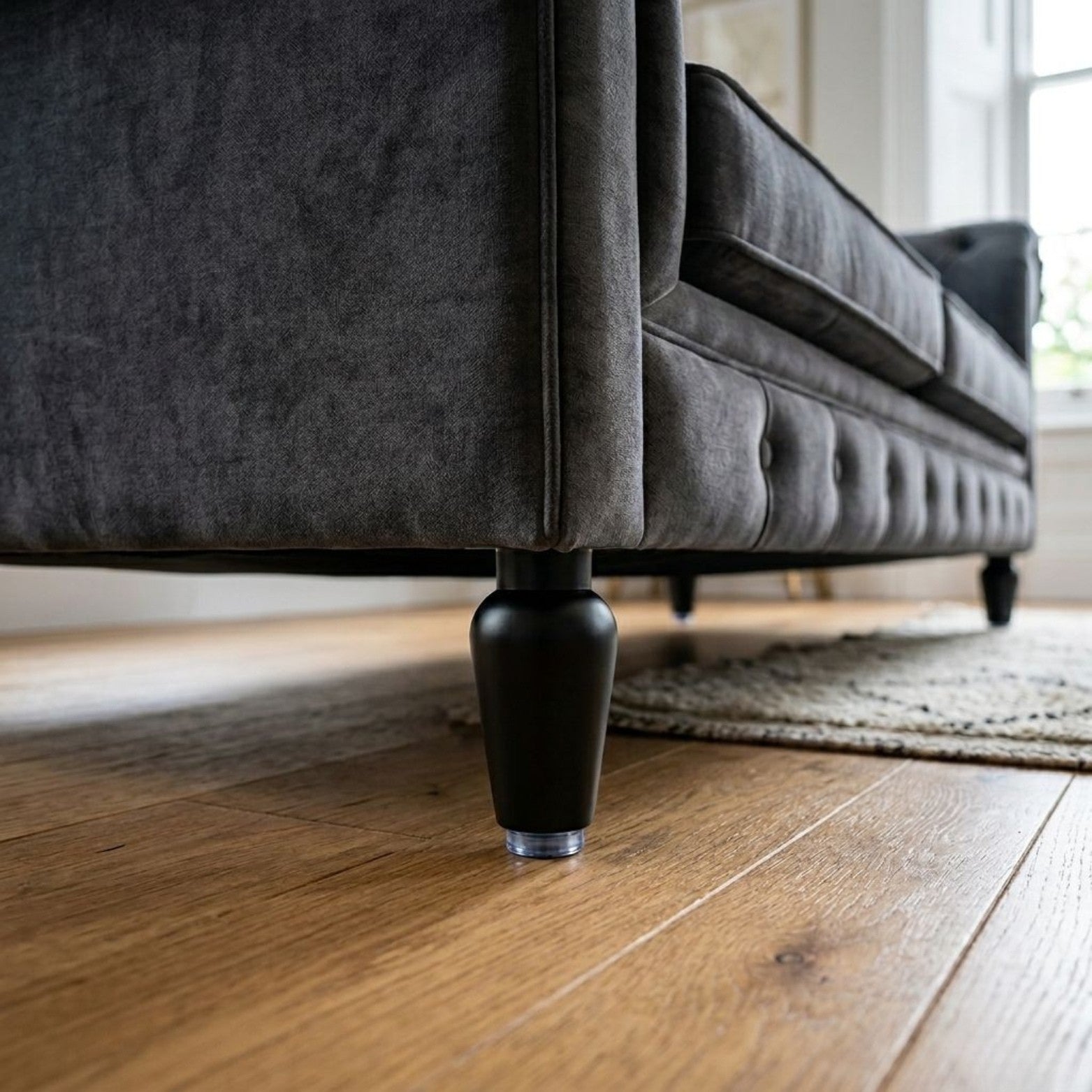 A sleek matte black metal sofa leg with a modern hourglass silhouette, featuring a clear protective floor glider on a wooden floor