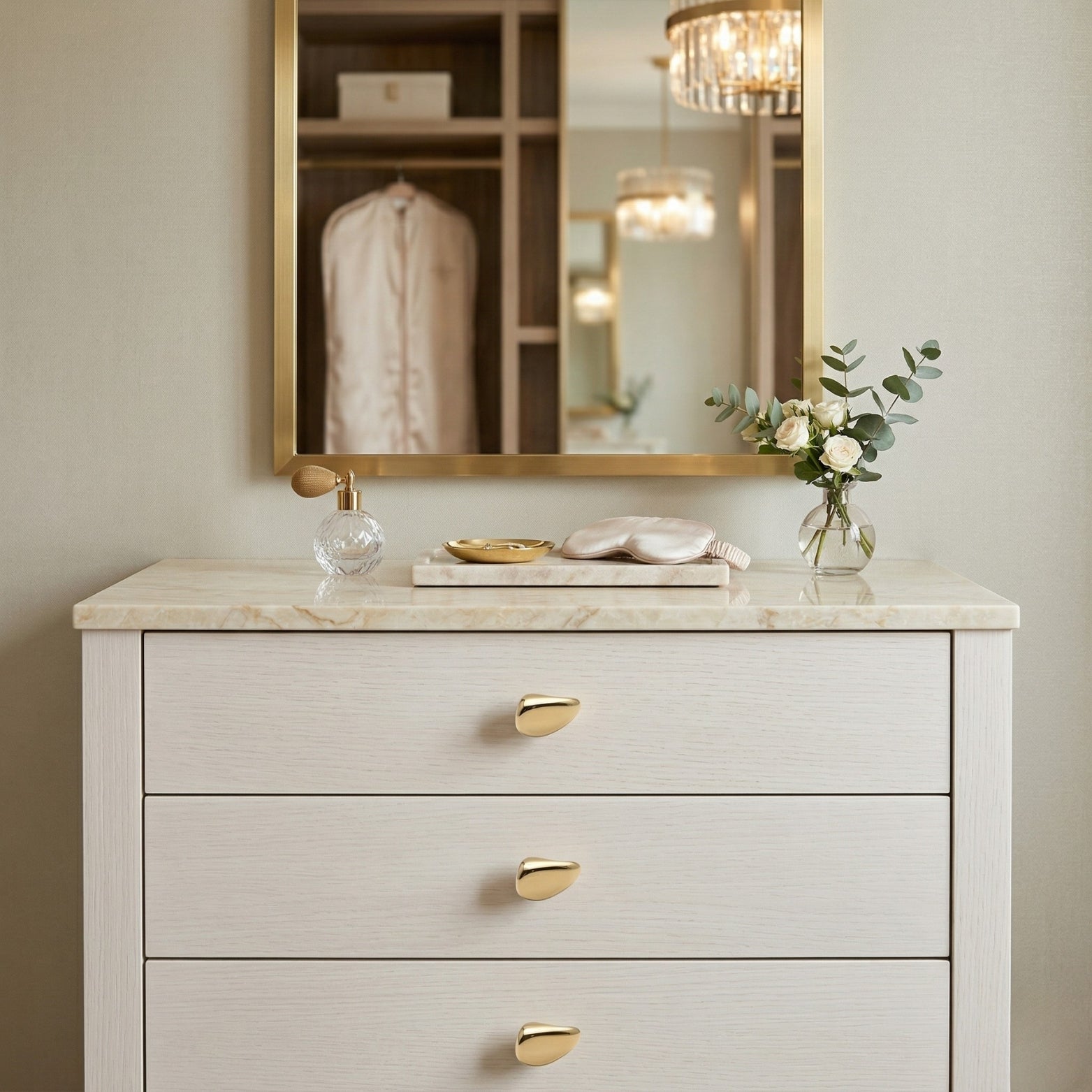 Close-up of a contemporary white wooden dresser featuring three polished brass, asymmetrical knob pulls with a distinctive pebble shape. The dresser is topped with marble, supporting a decorative arrangement including a vase of white roses and eucalyptus, a vintage perfume bottle, and a marble trinket dish. Above it hangs a brass-framed mirror reflecting a lit modern chandelier and a walk-in closet.