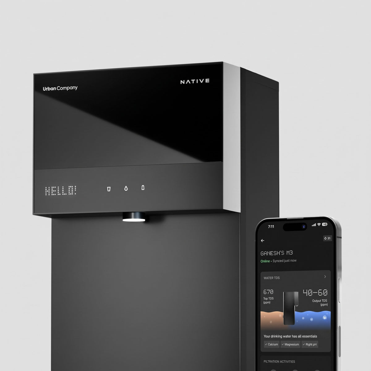A close-up of the black Urban Company Native water purifier alongside a smartphone in the foreground displaying the companion app, which highlights real-time input and output water TDS levels