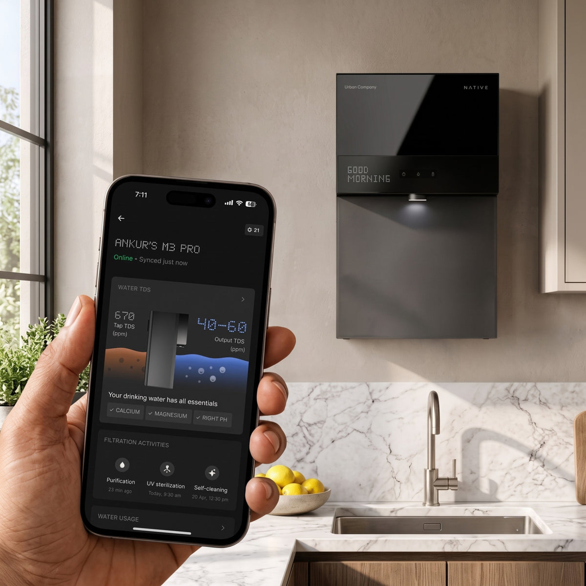 A hand holding a smartphone that displays the Native app dashboard showing TDS readings and filtration activities, with the black M3 Pro water purifier mounted on a bright kitchen wall in the background