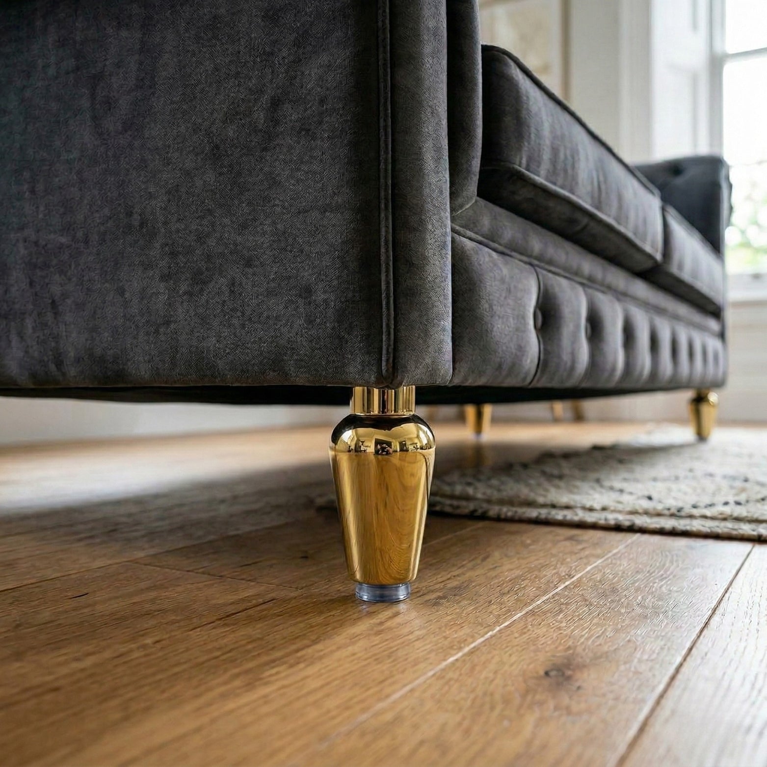 A luxurious polished gold or brass finish metal sofa leg in a tapered hourglass shape, attached to a dark grey tufted sofa