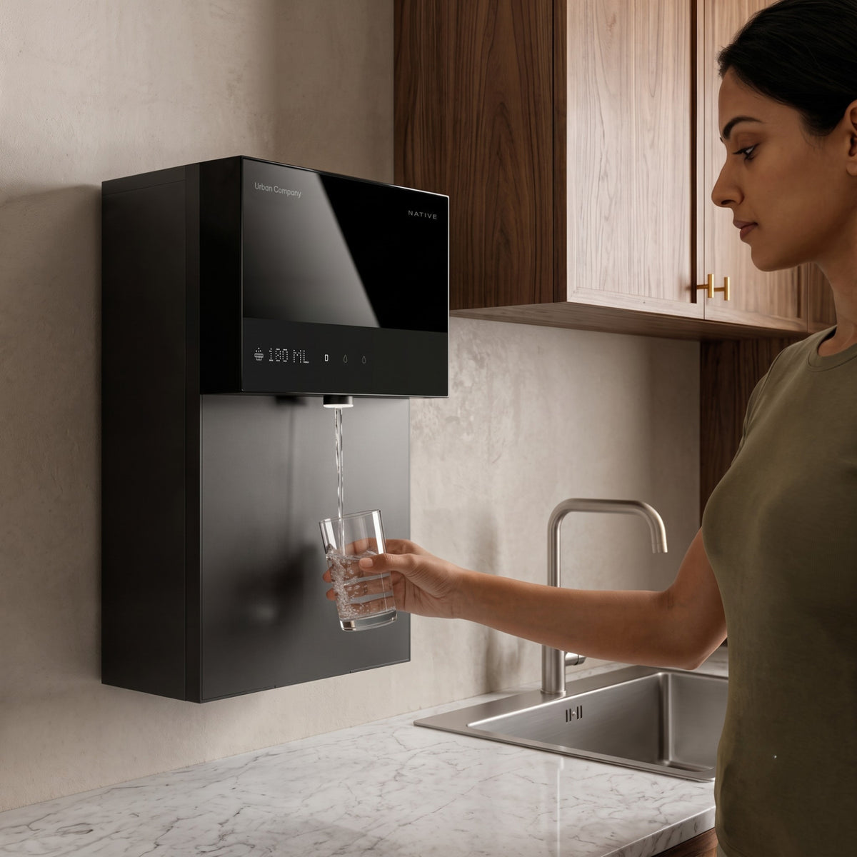 A woman standing in a modern kitchen filling a glass with water directly from the wall-mounted, black Urban Company Native M3 Pro water purifier, which displays the dispensing volume on its digital screen